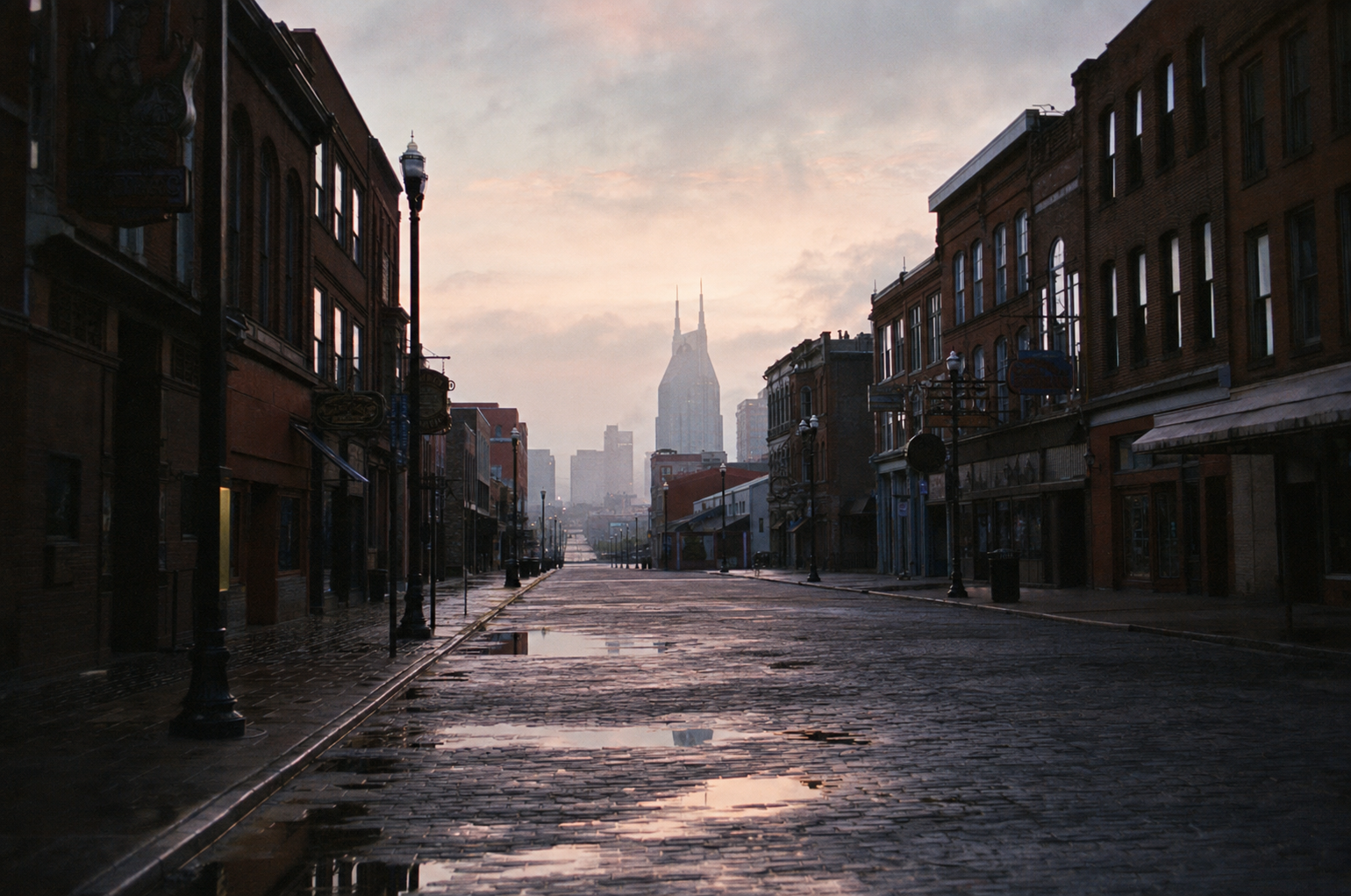 Lower Broadway Nashville at dawn, empty wet cobblestone street with historic brick honky-tonk facades and the AT&T tower silhouette in morning haze